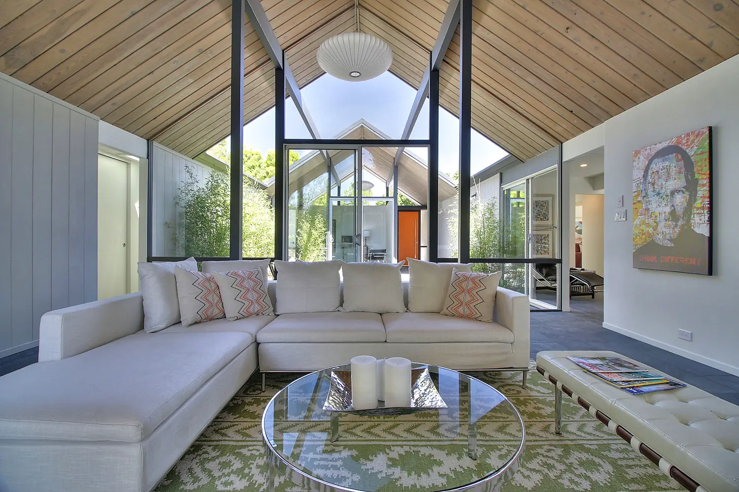 Looking across an Eichler living room through a full-height glass wall into the open-air atrium: exposed post-and-beam gable, tongue-and-groove ceiling, a Nelson bubble pendant, bamboo in the atrium and the orange front door beyond.