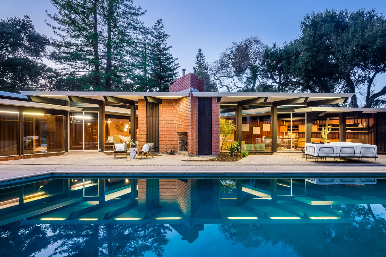 Joseph Eichler's own house at dusk, seen across the pool: a long post-and-beam elevation with deep overhangs, a central brick chimney mass and lit interiors under redwoods and oaks.
