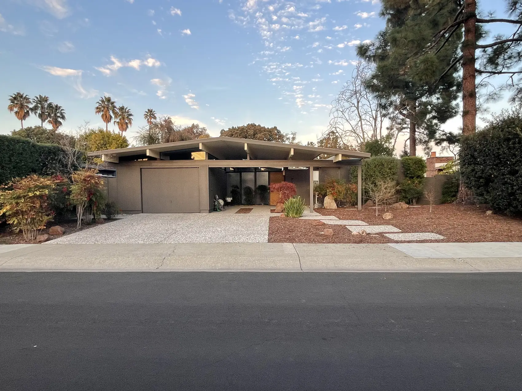 A Palo Alto Eichler at golden hour in the Los Arboles tract: low-slope gable roof with exposed beams, deep carport, aggregate driveway and mature palms.