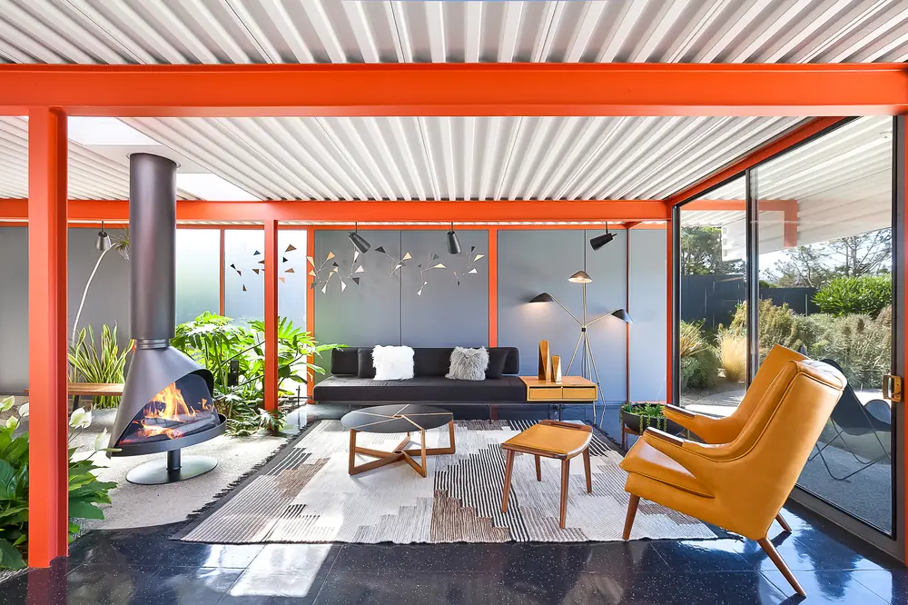 The X-100 living room: exposed orange steel columns, a corrugated metal ceiling, a suspended conical fireplace and a full glass wall onto the garden.