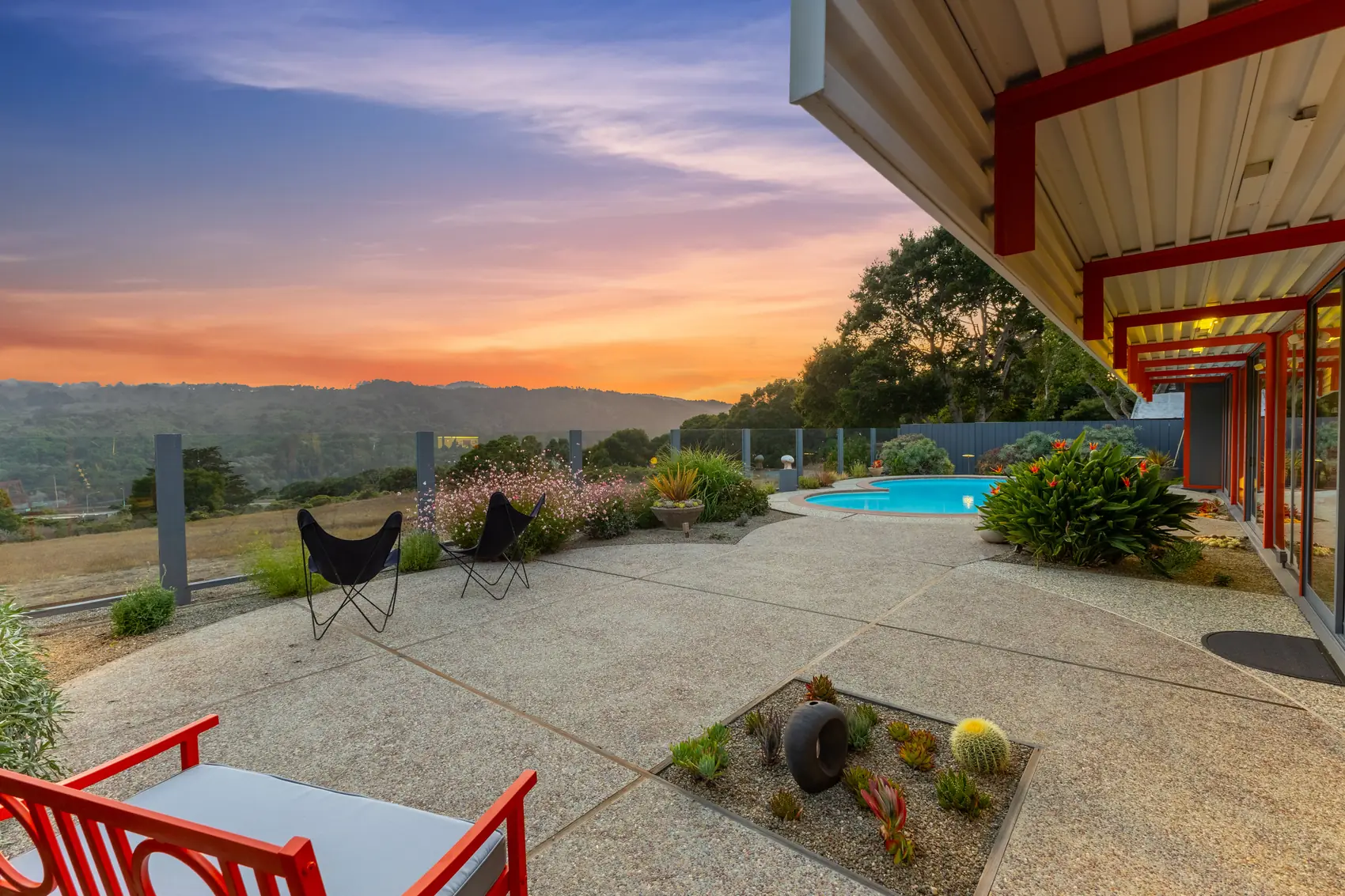 The Eichler X-100 at sunset: orange steel beams over a concrete terrace, a circular pool, butterfly chairs and the San Mateo hills beyond.